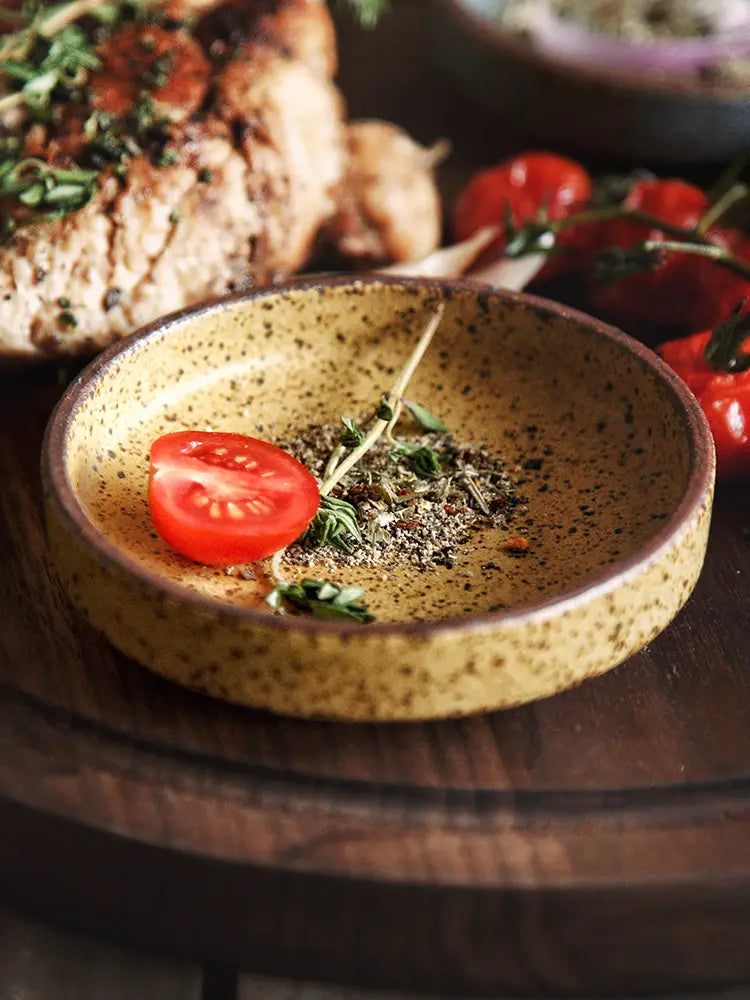A speckled yellow ceramic sauce dish rustic Japanese dipping bowl holding a halved cherry tomato and fresh herbs.