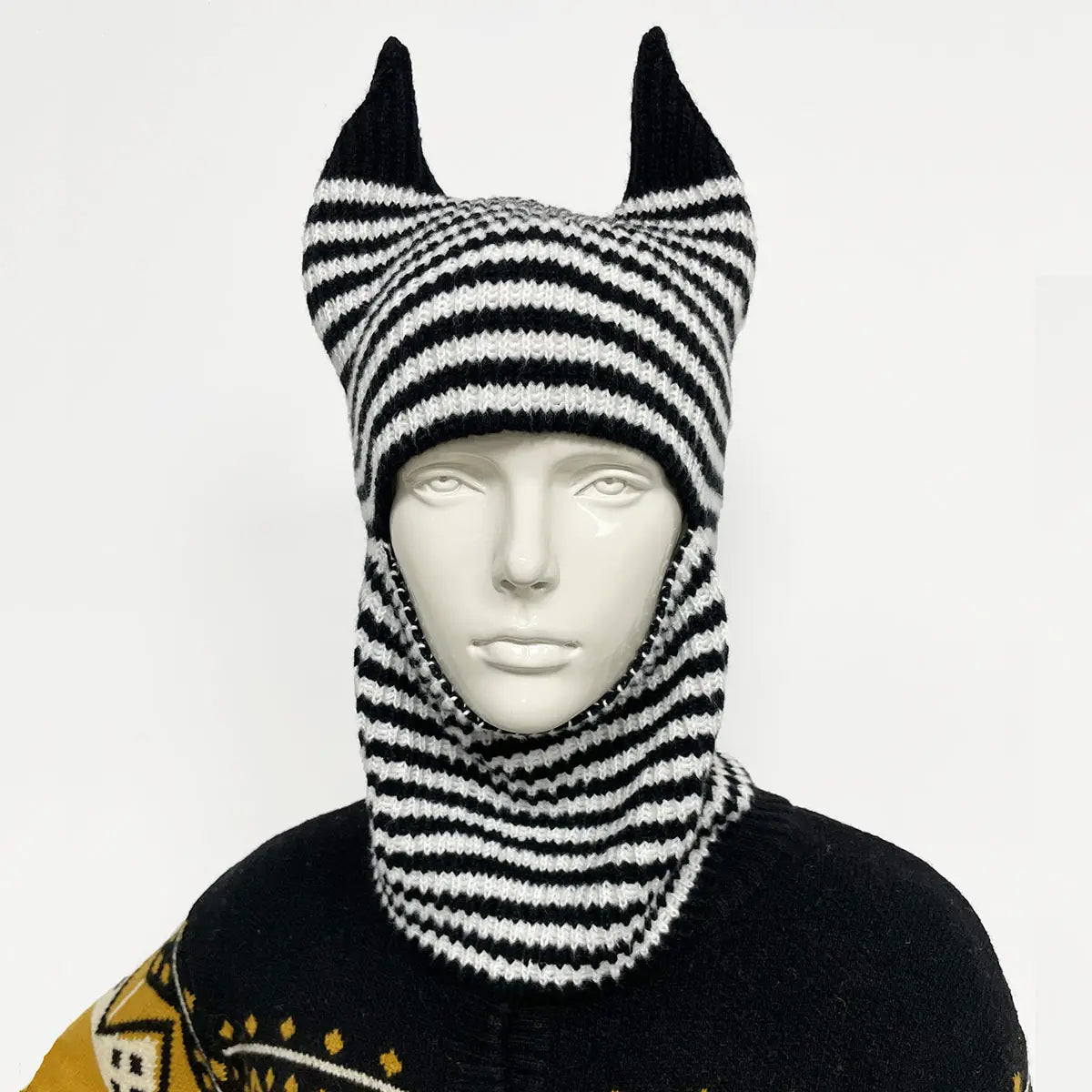 Bonnet hivernal de type cagoule tricoté à rayures noir et blanc, présentant des protubérances pointues en forme d’oreilles et une texture tricotée.