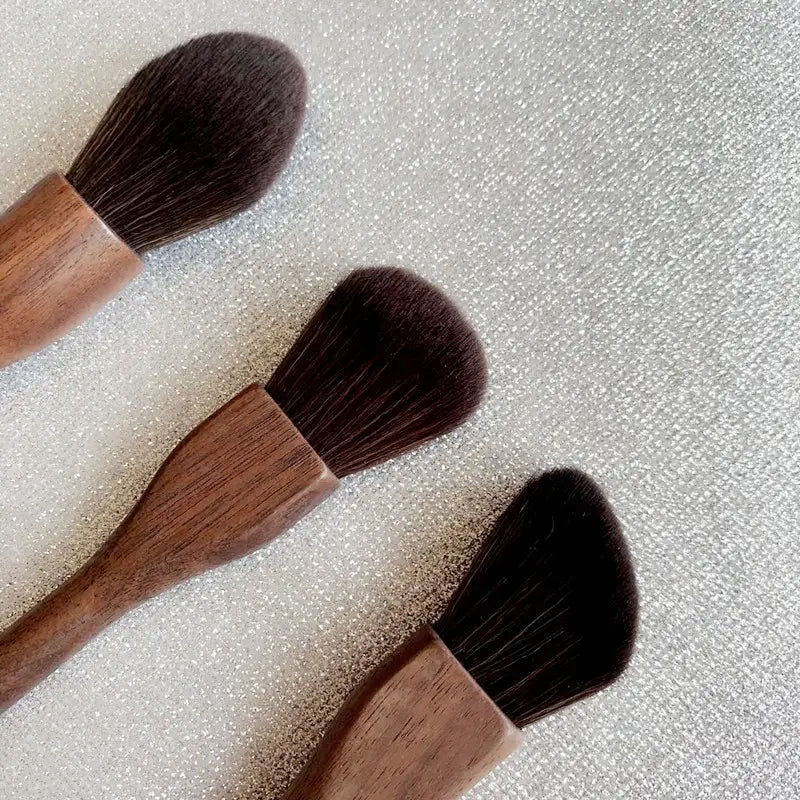 Three wooden-handled walnut wood makeup brushes with soft dense dark brown fiber bristles arranged diagonally on a textured silver surface.
