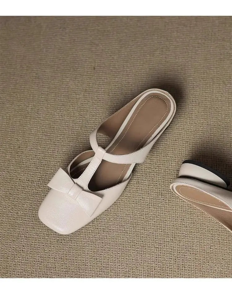 Cream-colored closed toe leather mule flats for women with a square toe, glossy finish, and decorative bow on the front.