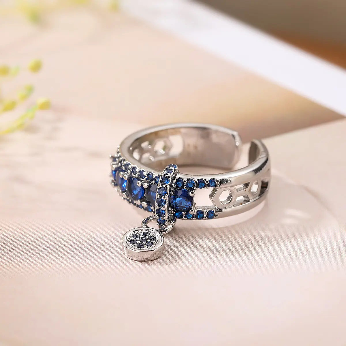 Adjustable geometric silver open ring adorned with blue zircon gemstones and a dangling circular charm featuring black stones.