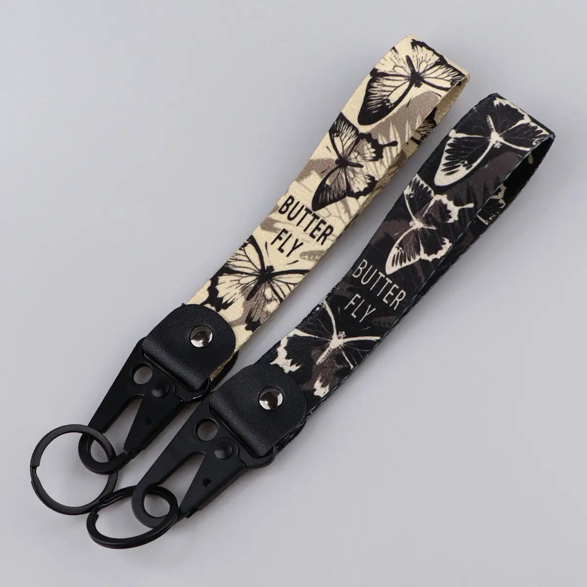 Two butterfly wrist strap keychains with metal clips in beige with black butterfly patterns and black with white butterfly patterns featuring fabric straps labeled butterfly attached to black metal carabiners.