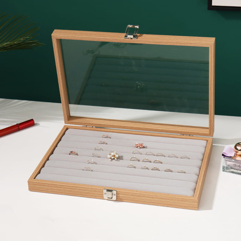 Rosewood jewelry display box with light wood finish rectangular shape clear glass lid silver toned metal clasps and padded gray interior dividers holding various rings.