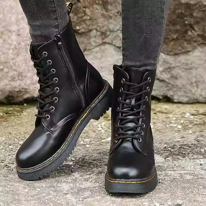 Botines estilo Martin de cuero negro con punta redonda, parte frontal con cordones, suela de goma gruesa y costuras amarillas, diseñados como calzado plano a la altura del tobillo.