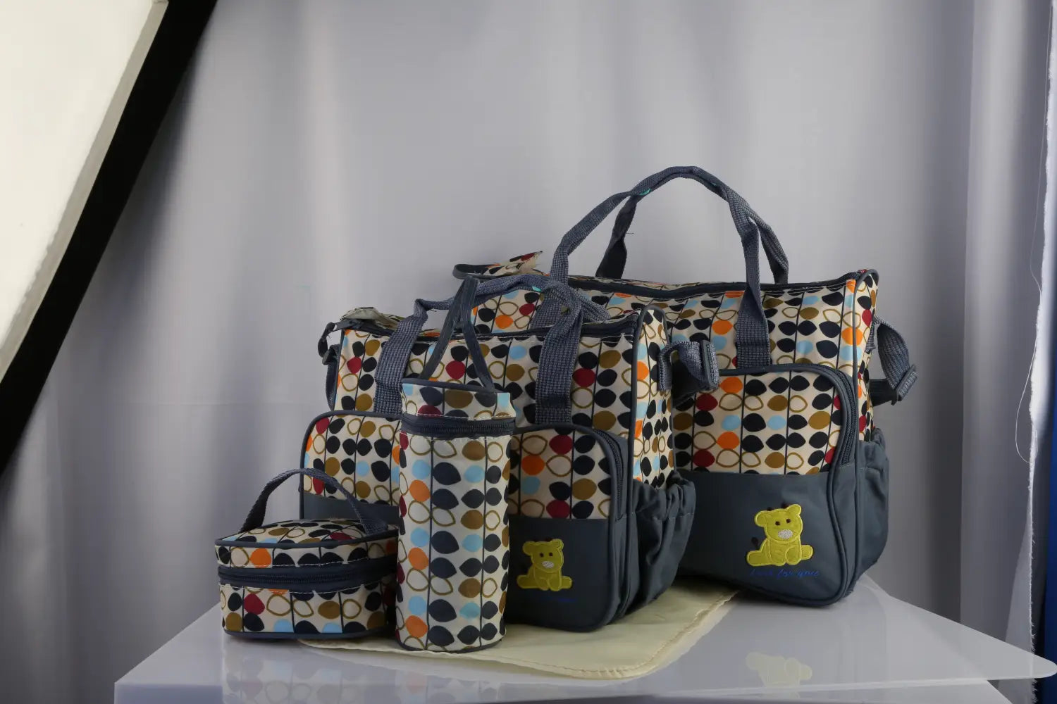 Navy blue and multicolored polka dot 5-piece floral water-resistant baby diaper bag travel set including a large tote, smaller zippered pouch, and cylindrical bottle holder with yellow bear appliqués.