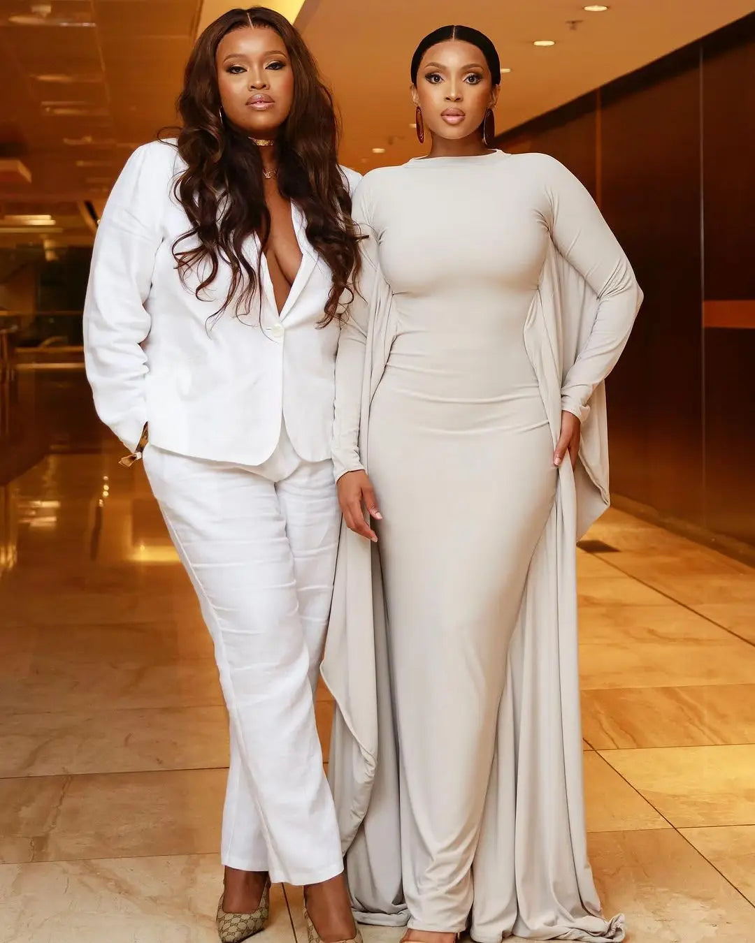 The woman on the right wears an off-white asymmetrical satin batwing sleeve maxi dress with long sleeves and a draped cape-like overlay.