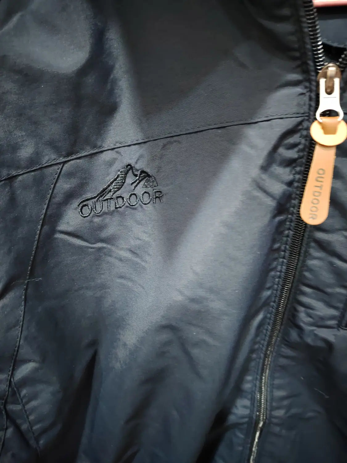 Chaqueta de senderismo con capucha impermeable color negro, con un logotipo de montaña sutil bordado en el pecho y un tirador de cremallera de cuero color camel con la etiqueta “outdoor”.