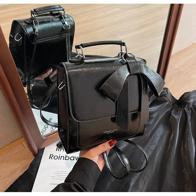 Schwarze, glänzende Leder-Umhängetasche mit auffälliger Schleifendetailierung und Metallbeschlägen für den Einsatz als modische Handtasche.
