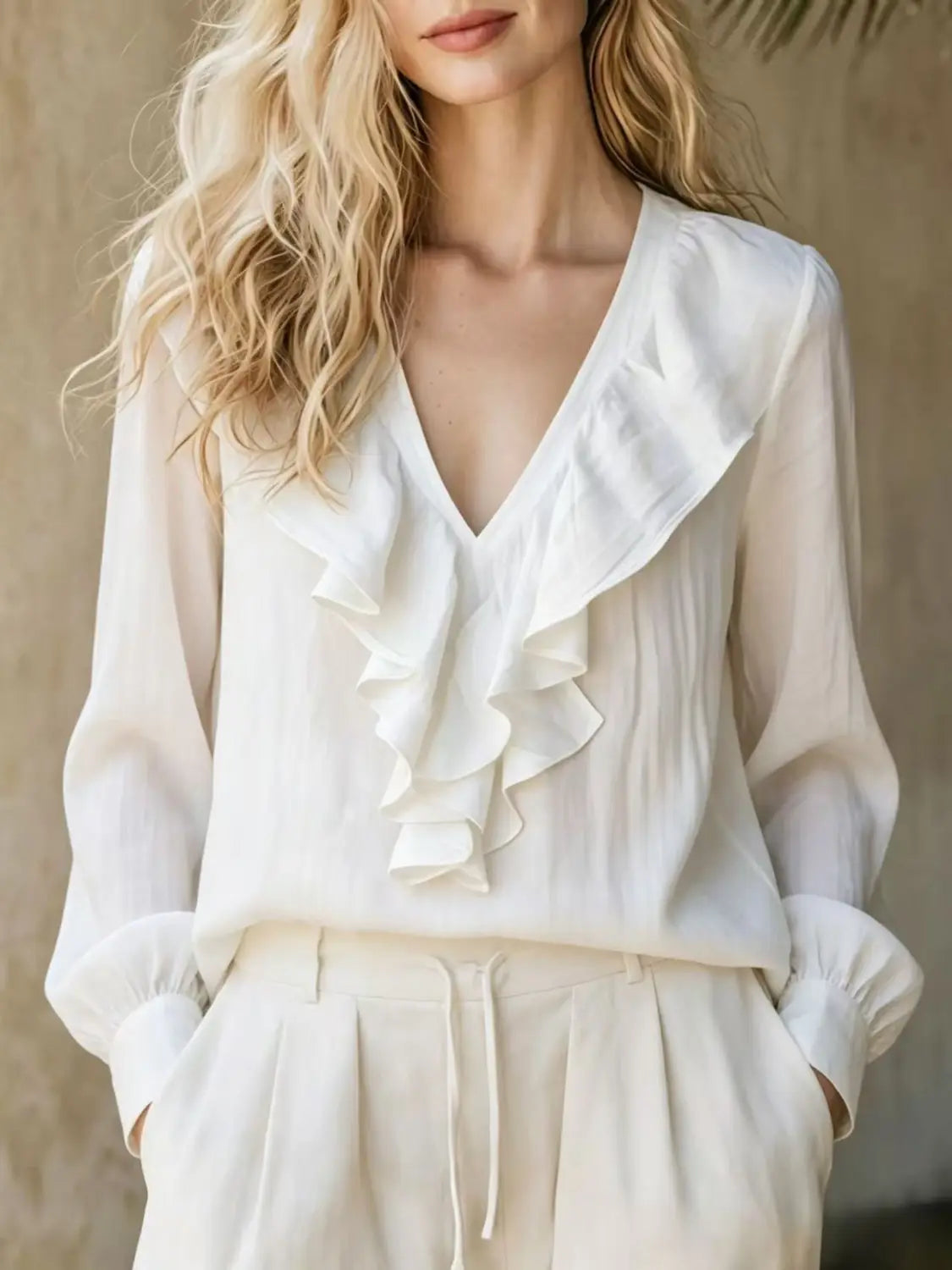 Une blouse en lin blanche fluide à encolure en V et manches longues, avec des détails de volants délicats le long de l’encolure et des manches.