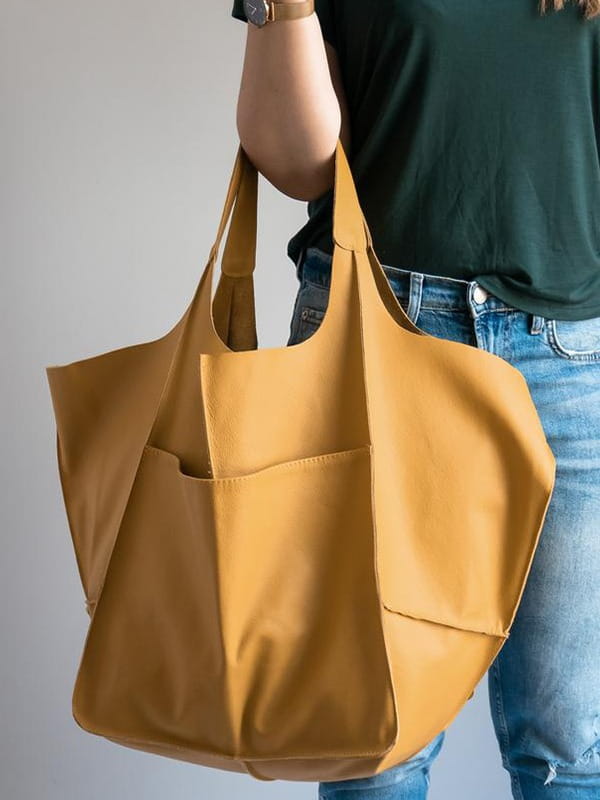 A mustard-yellow leather tote bag with a large capacity, soft slouchy shoulder design, structured geometric shape, and two front pockets.