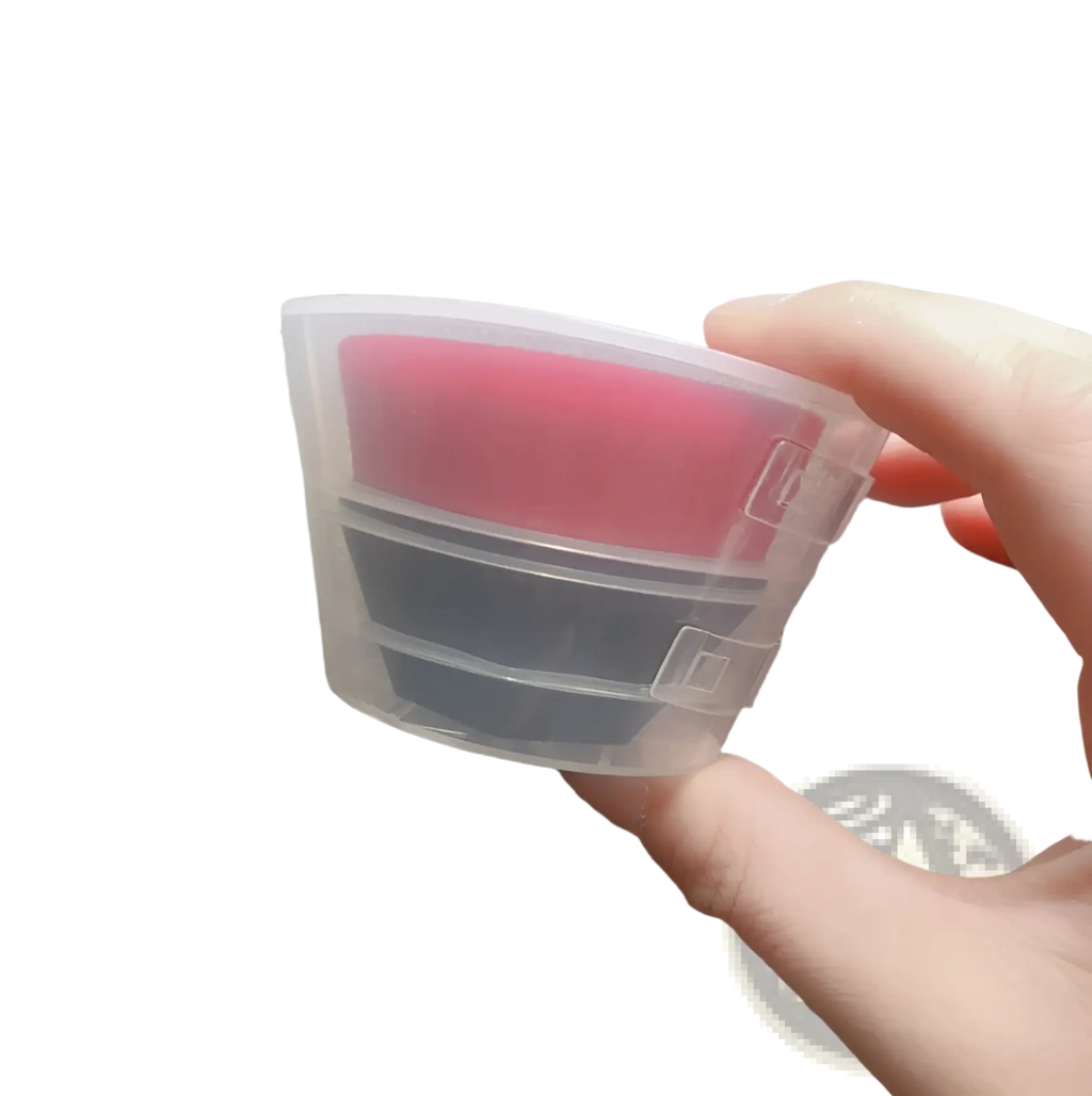 Transparent plastic container holding two stacked silicone pads in pink and dark gray for foundation brush and powder set