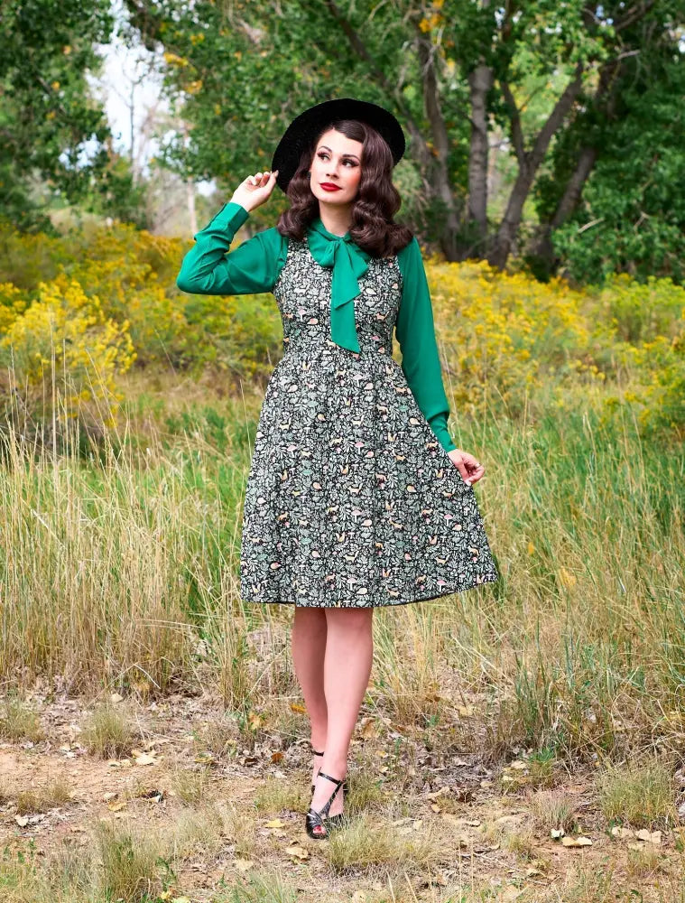 A woman wears a foxy vintage green fit and flare nature print dress with a green long-sleeved top and black hat