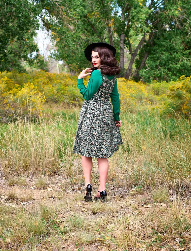 A woman wears a foxy vintage green fit and flare nature print dress with long sleeves and a wide-brimmed black hat