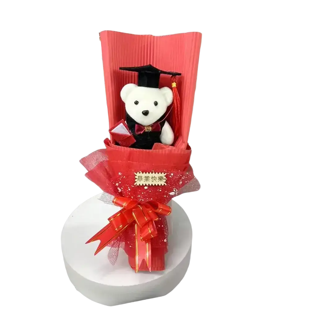 A plush white graduation bear wearing a black cap and gown with red bow tie in a red floral bear bouquet