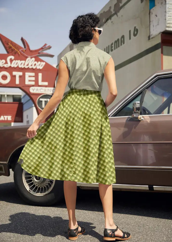 A woman wears a sleeveless olive green and white checkered cotton a-line skirt with fitted waistband and pockets