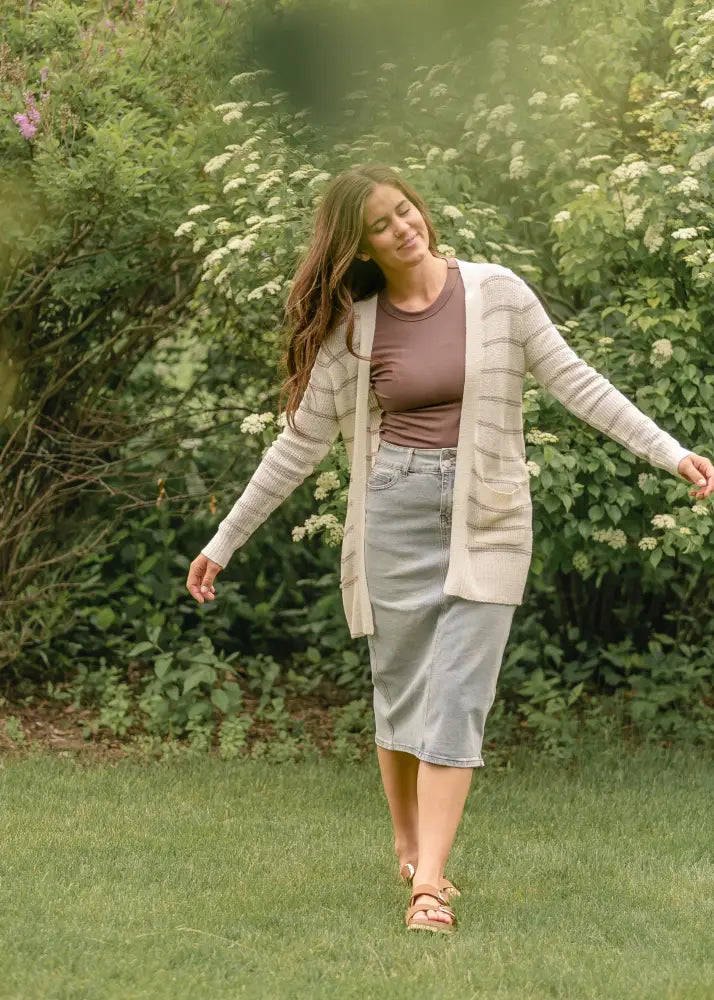 A woman wearing a beige cardigan with horizontal stripes over a brown top and a Greta light wash button front denim midi skirt