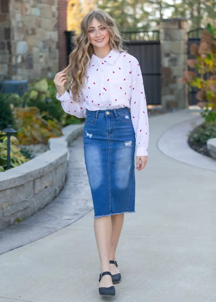 A woman wearing a white blouse with red heart patterns paired with a hadley high rise distressed denim midi skirt