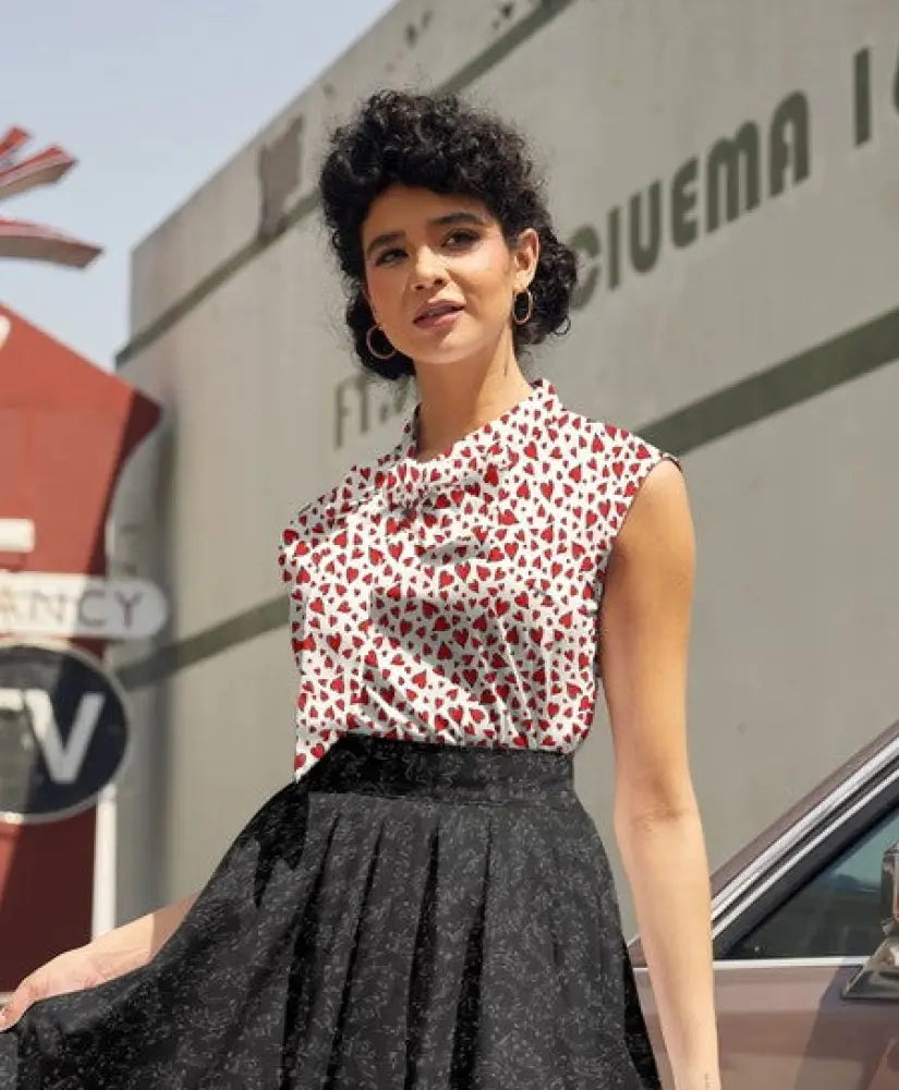 A woman wears a vintage 1950s white sleeveless heart print bow top with red hearts paired with a black pleated skirt
