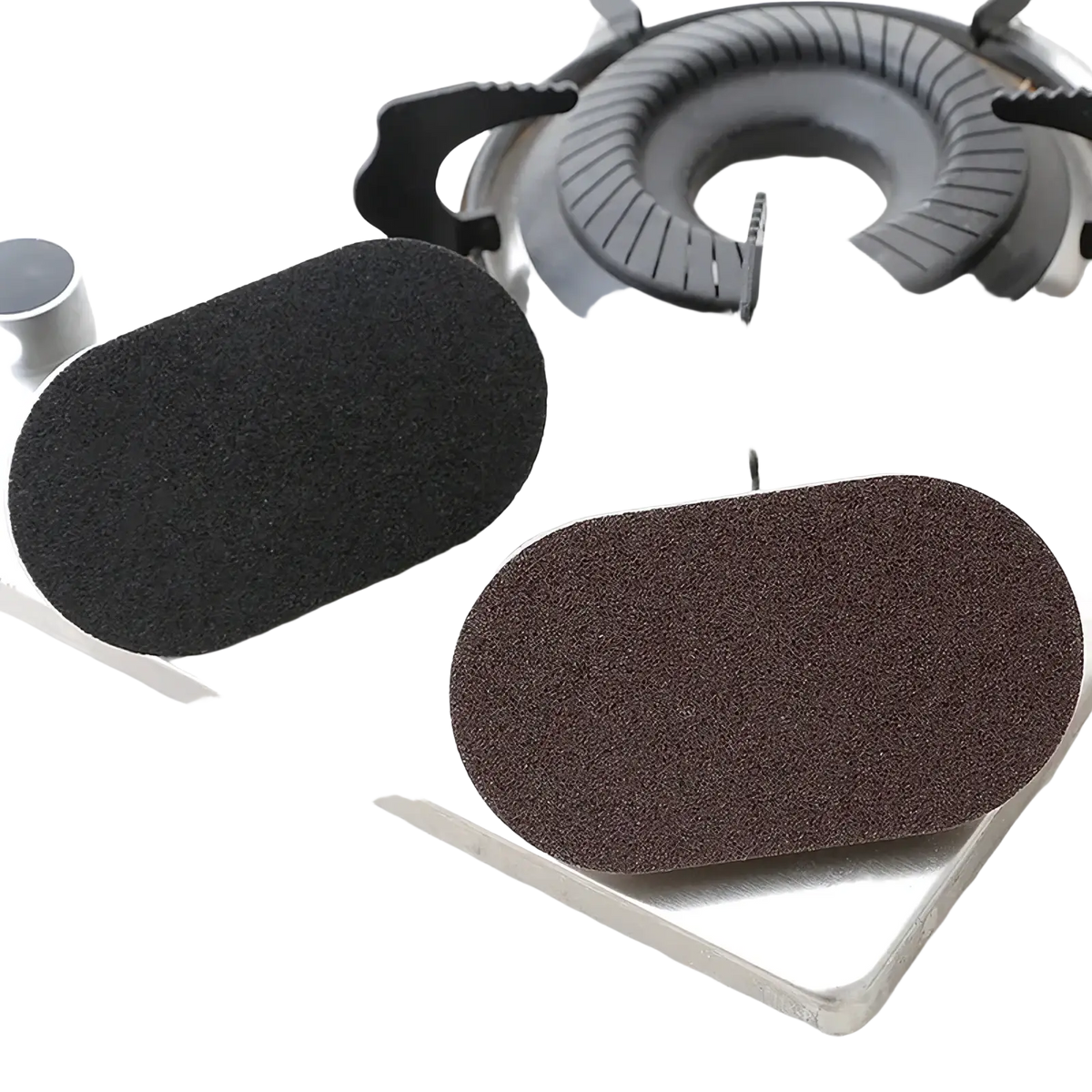 Two oval heavy duty pot scraper sponges in black and brown with textured surfaces near a gas stove burner