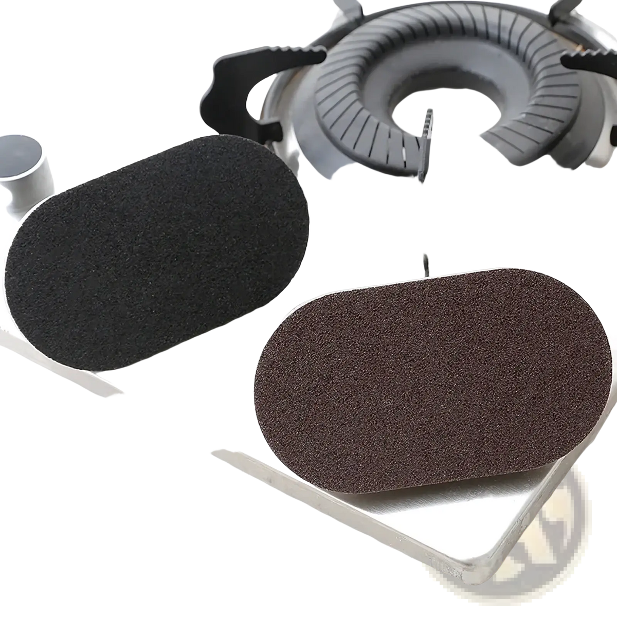 Two oval heavy duty pot scraper sponges in black and brown with textured surfaces near a gas stove burner