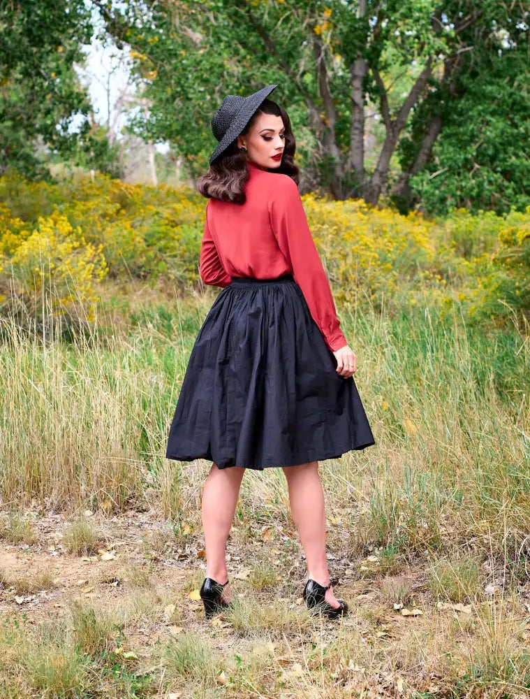 A woman wears a helen retro bow tie blouse in cognac with long sleeves paired with a black full skirt and wide-brimmed hat
