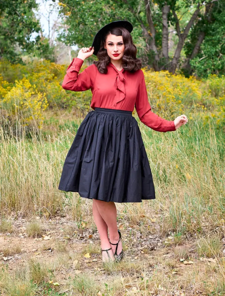 A woman wears a helen retro bow tie blouse in cognac with a ruffled neckline and a black pleated skirt