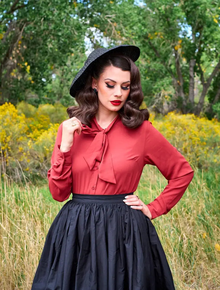 A woman wearing a helen retro bow tie blouse in cognac red with a bow neckline paired with a black skirt and hat