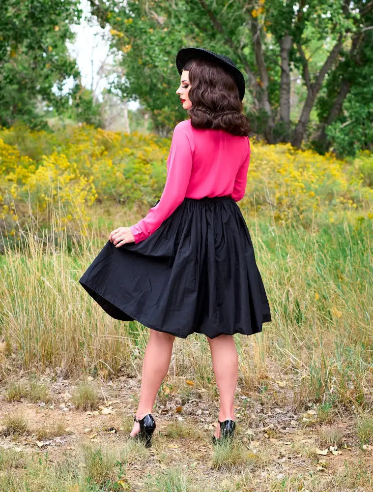 A woman wearing a helen pink bow tie retro long sleeve blouse paired with a black a-line skirt and black heels