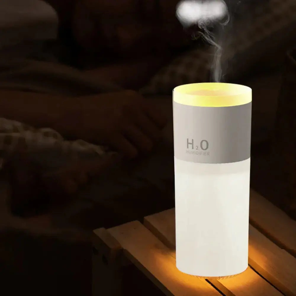 A sleek cylindrical white and gray portable usb led mist humidifier and aroma diffuser with a glowing yellow top emitting mist