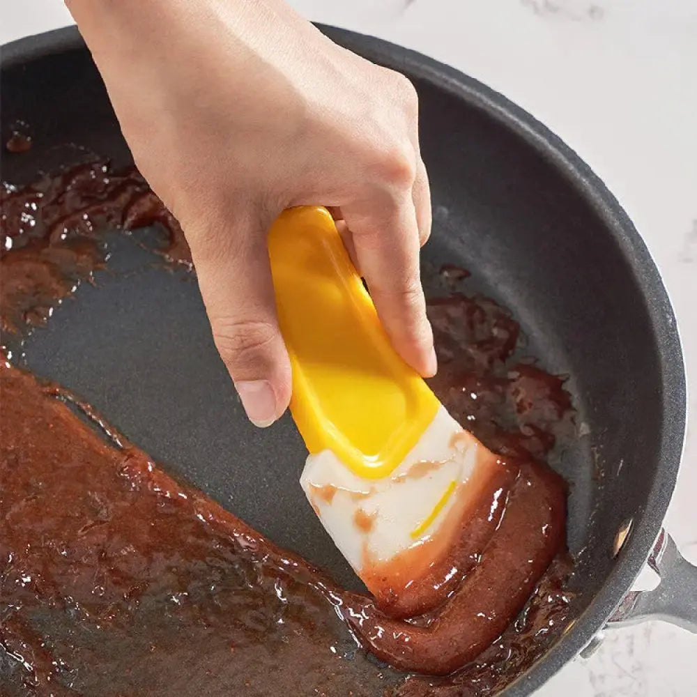 Yellow silicone kitchen squeegee with a white blade scraping sauce from a black frying pan in use