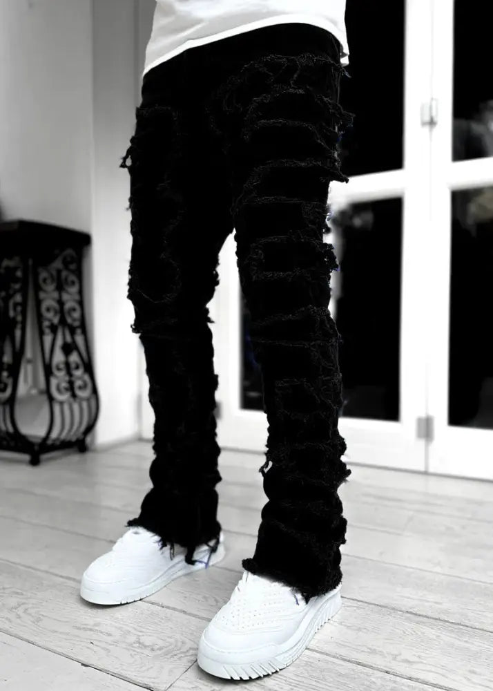 Men’s slim fit stacked ripped patchwork streetwear jeans in black distressed denim with frayed ripped detailing along legs