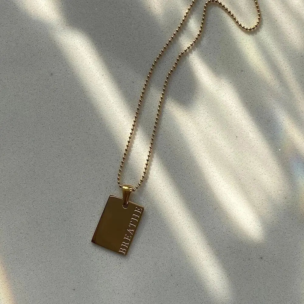 Gold layered rectangular pendant tag necklace with breathe engraved on the side suspended from a delicate ball chain