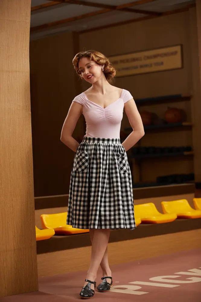 A woman wears an Isabel ruched sweetheart top in blush pink paired with a black-and-white gingham skirt with pockets
