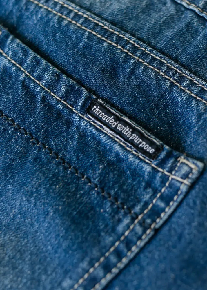 A dark blue denim pocket with white stitching and a black label reading threaded with purpose on the back of a jennifer premium a-line denim maxi skirt with flared hem