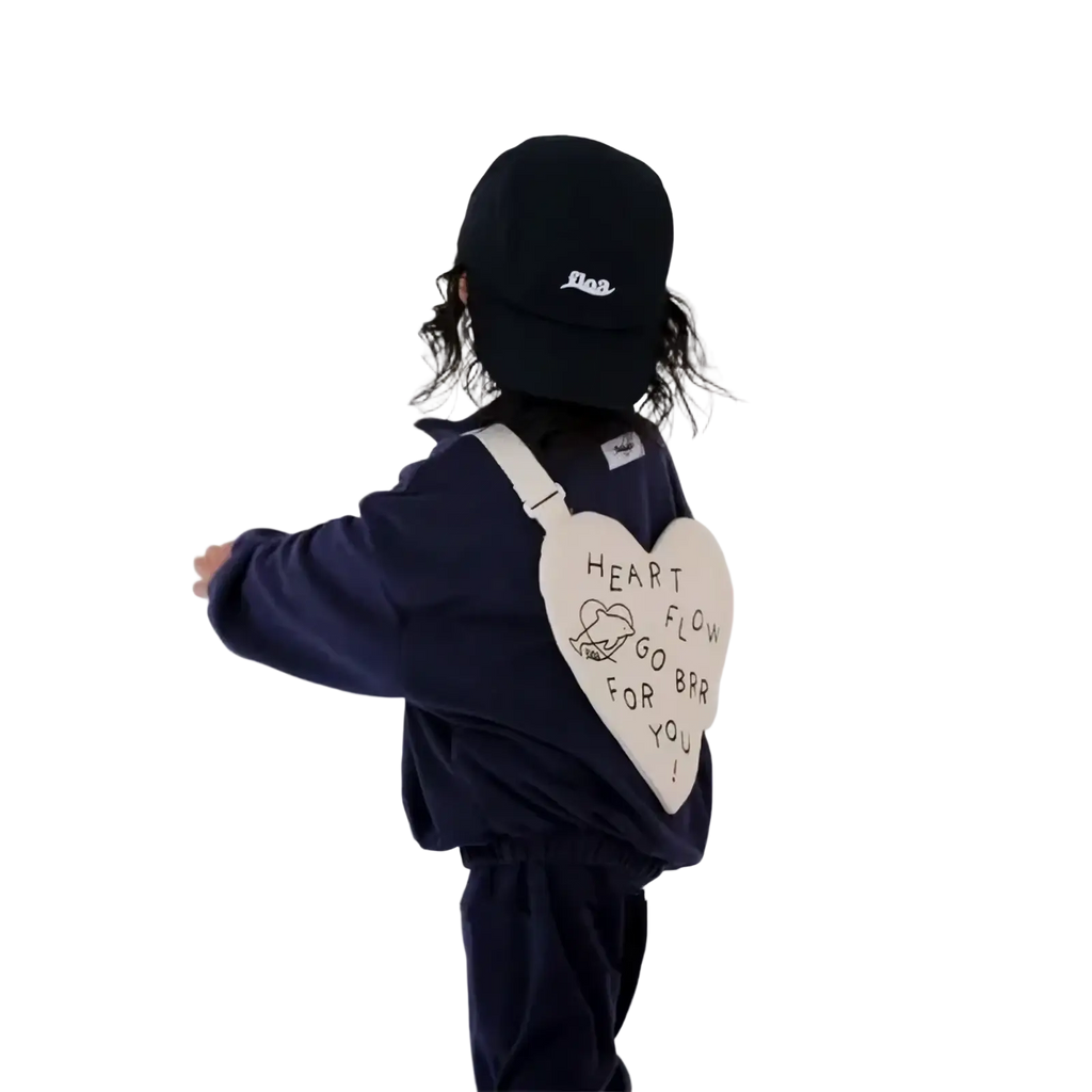 A cream-colored heart-shaped Korean kids crossbody bag with black embroidery worn by a child in navy blue clothing