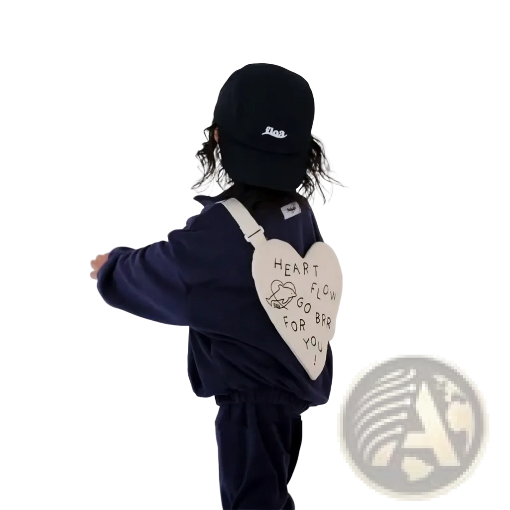 A cream-colored heart-shaped Korean kids crossbody bag with black embroidery worn by a child in navy blue clothing