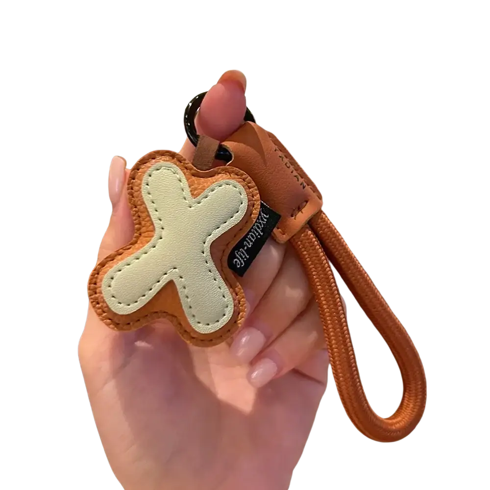 A tan leather initial keychain wristband charm with cream cross-shaped detail and an orange woven strap