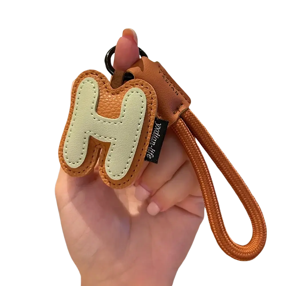 A tan leather initial keychain with a light beige stitched letter h and an orange braided wristband charm