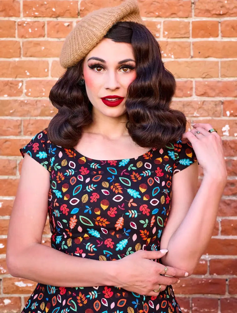 A woman wears a black cotton Margaret vintage a line dress with colorful stylized autumn leaves and acorns