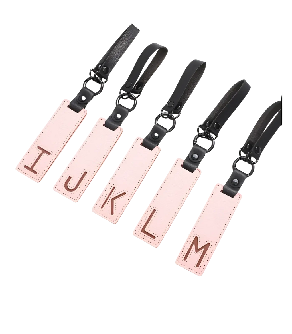 Pink leather luggage tags with black straps and metal rings featuring brown embossed uppercase letters for alphabet keychains