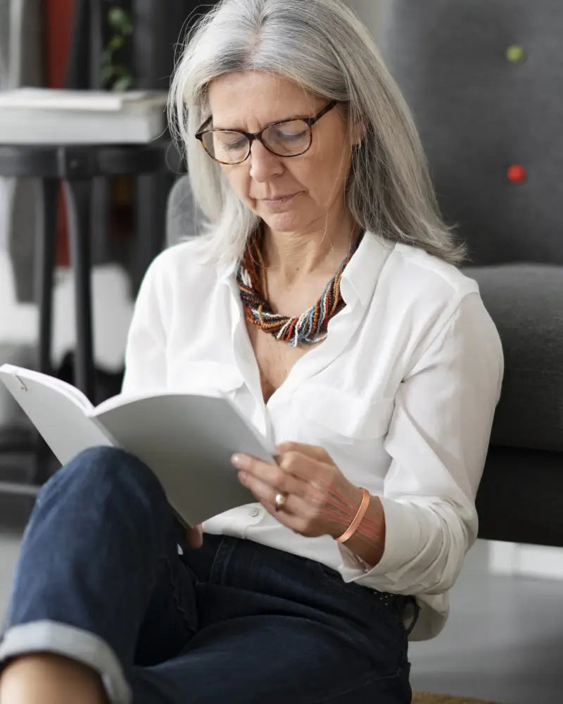 A woman with silver hair and glasses reads a book while wearing a rose gold pure copper magnetic therapy cuff bracelet