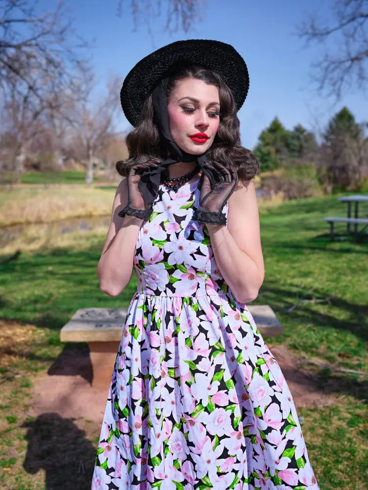 A woman wears a black straw hat and a magnolia floral cotton 1950s style midi dress with pink blossoms and pockets