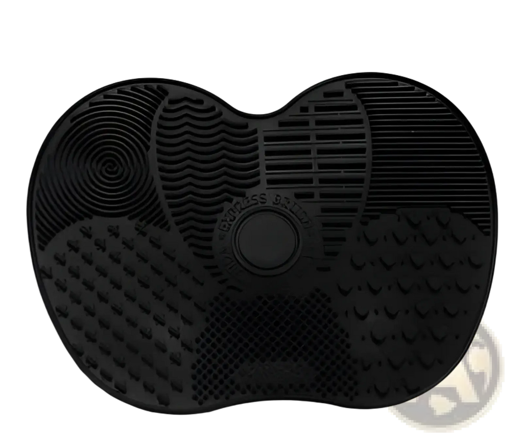Black silicone heart shaped grooming mat with textured surfaces and central circular button for makeup brush cleaning