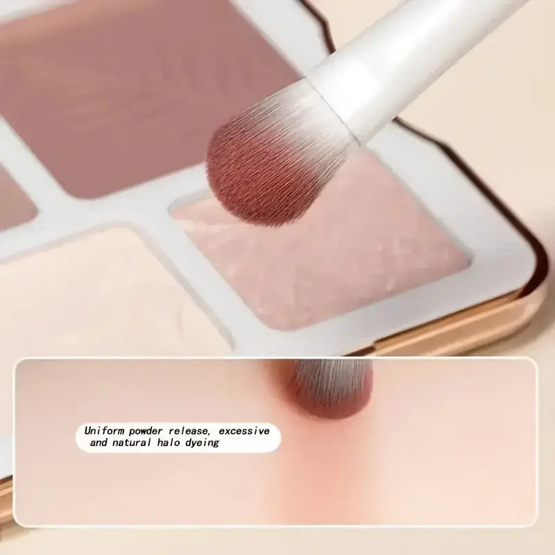 A white-handled makeup brush with soft tapered bristles gently applies light pink powder from an open eyeshadow palette