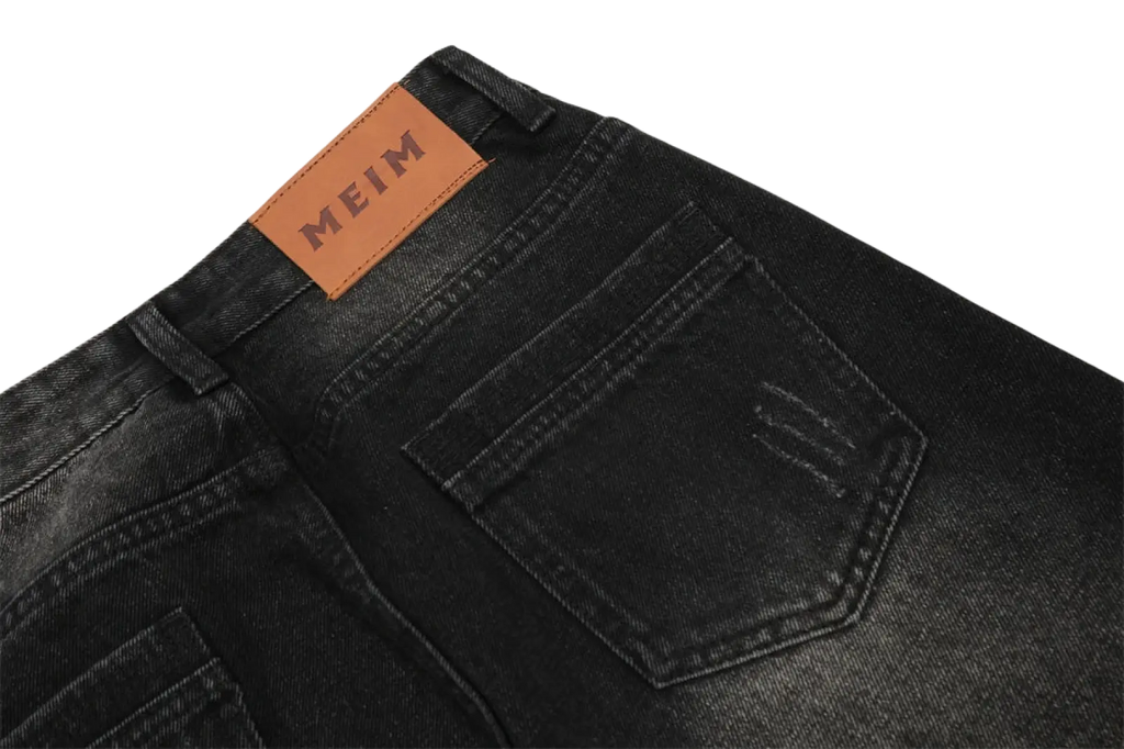 Back side of men’s distressed straight leg black denim jeans featuring a brown leather meim brand patch embossed in black lettering