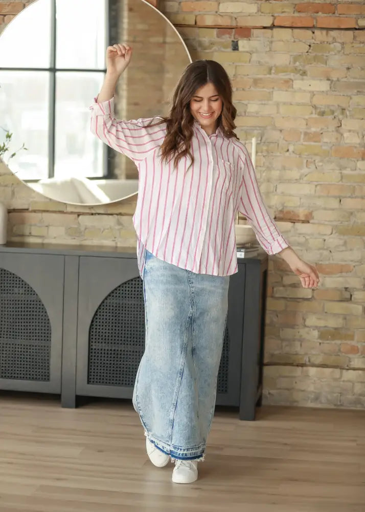 A woman wears a mina frayed hem light wash denim maxi skirt paired with a pink and white striped button-up shirt