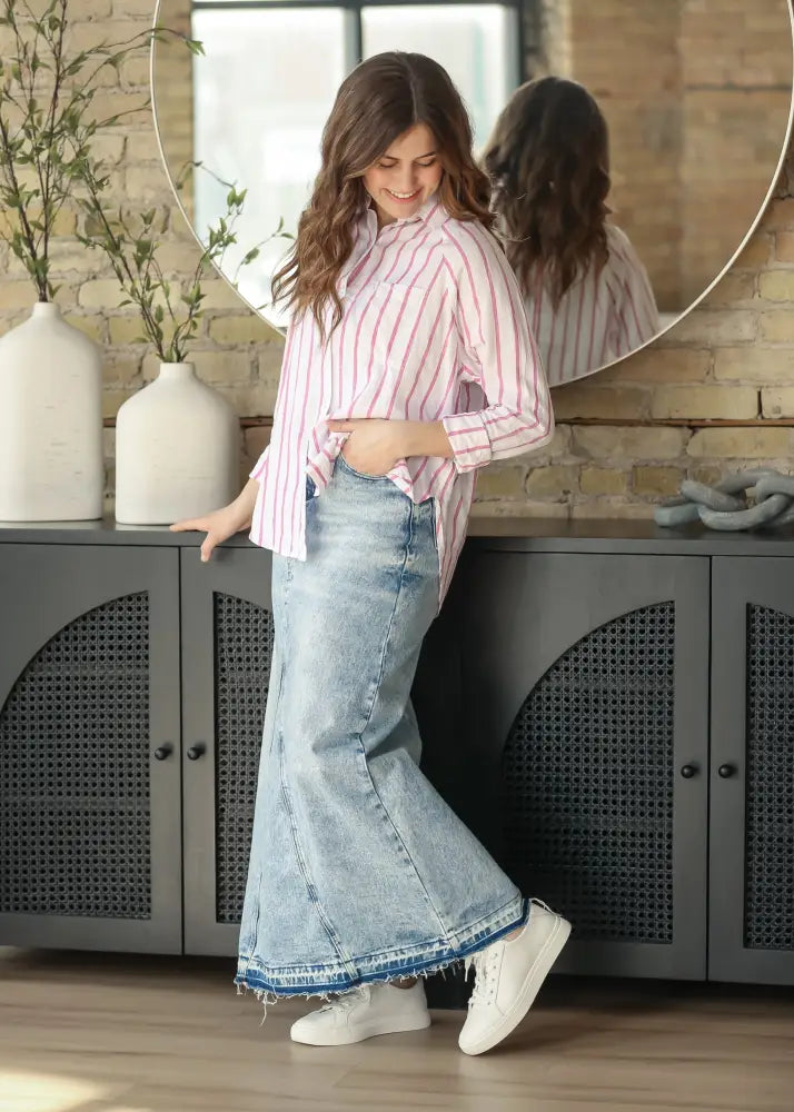 A woman wearing a pink and white striped button-up shirt with a mina frayed hem light wash denim maxi skirt