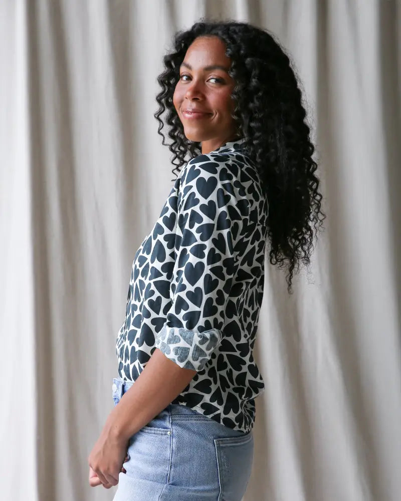 A woman wearing a white cotton More Love Giraffe heart print button up blouse with black hearts and rolled sleeves