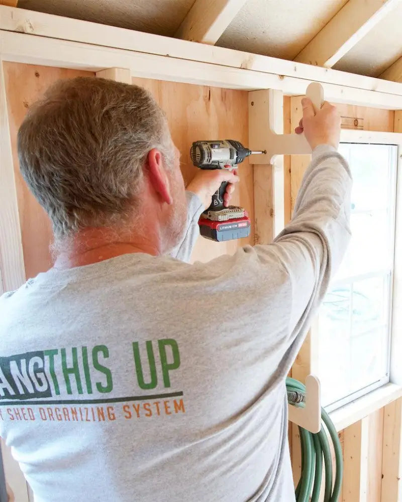 A man uses a black and red cordless drill to attach engraved wooden stud mounted utility storage hooks inside a shed