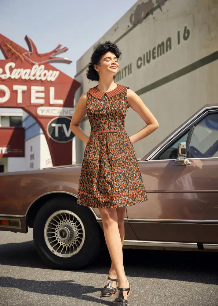 A woman wears a retro 60s mushroom print a-line cotton dress with pockets in brown orange and black geometric pattern